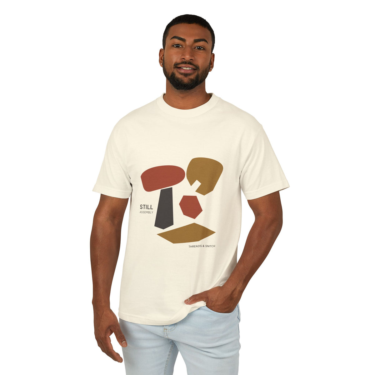 Model mockup of Still Assembly Abstract Graphic Tee in Faded Cream, front view showing floating rounded and angular sculptural forms with Still Assembly lettering.