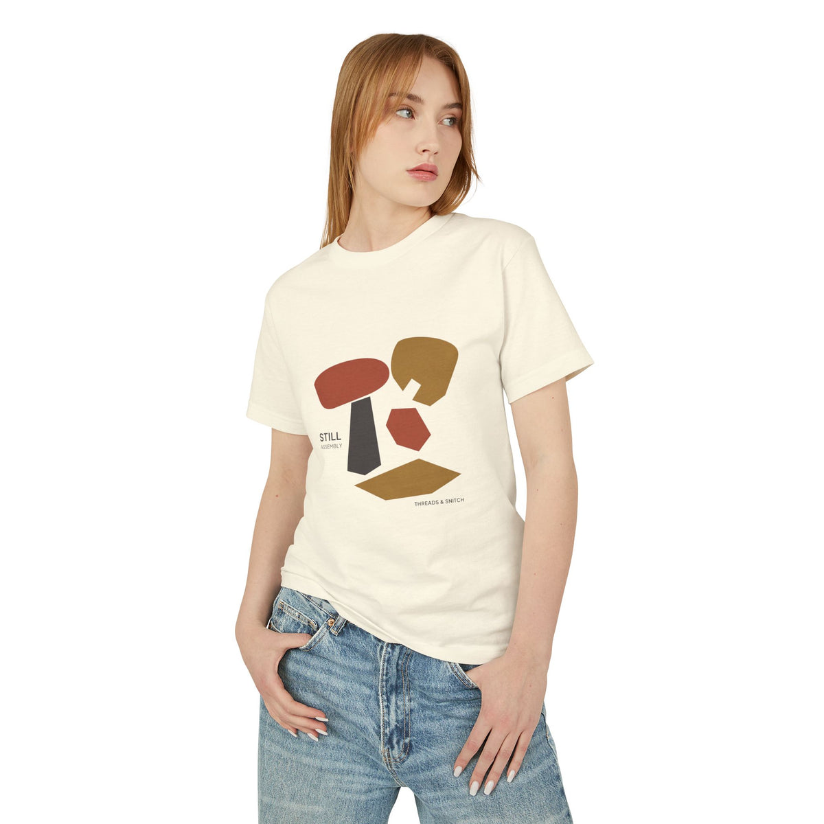 Model mockup of Still Assembly Abstract Graphic Tee in Faded Cream, front view showing floating rounded and angular sculptural forms with Still Assembly lettering.