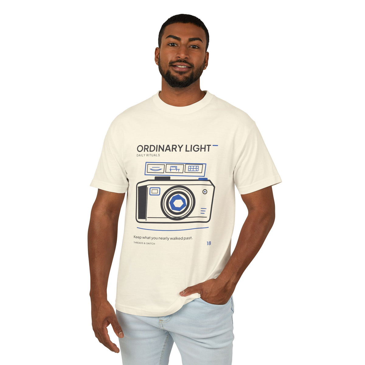 Model mockup of Ordinary Light Camera Tee in Faded Cream, front view showing camera with blue lens details and a strip of small domestic scenes.