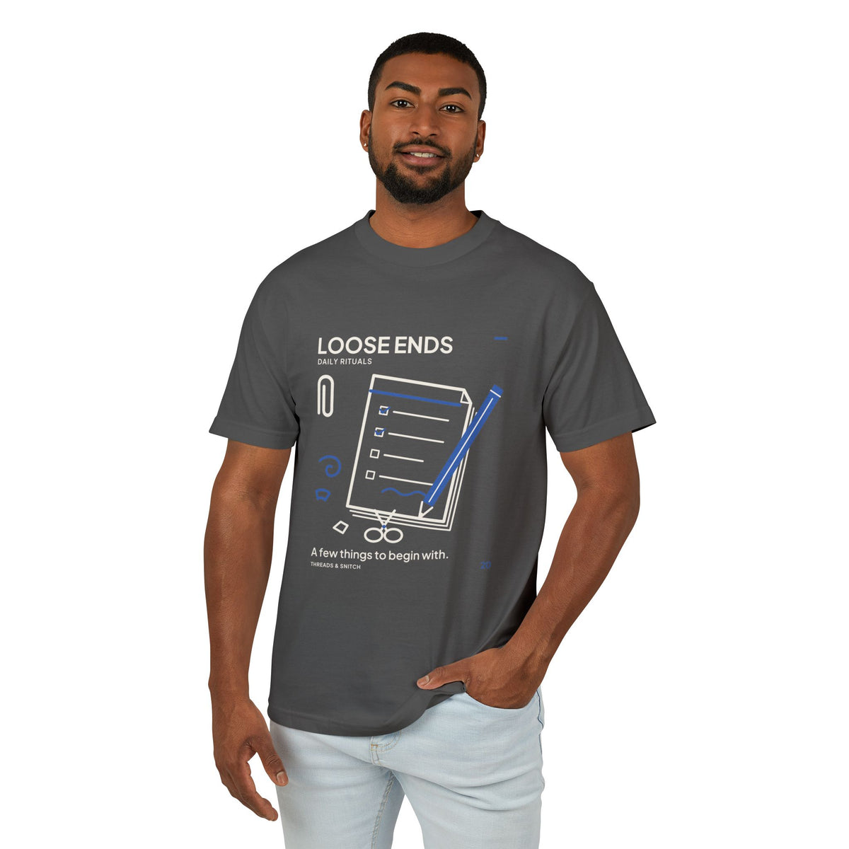 Model mockup of Loose Ends Stationery Tee in Faded Black, front view showing diagonal pencil over a task pad with scissors and a paper clip.