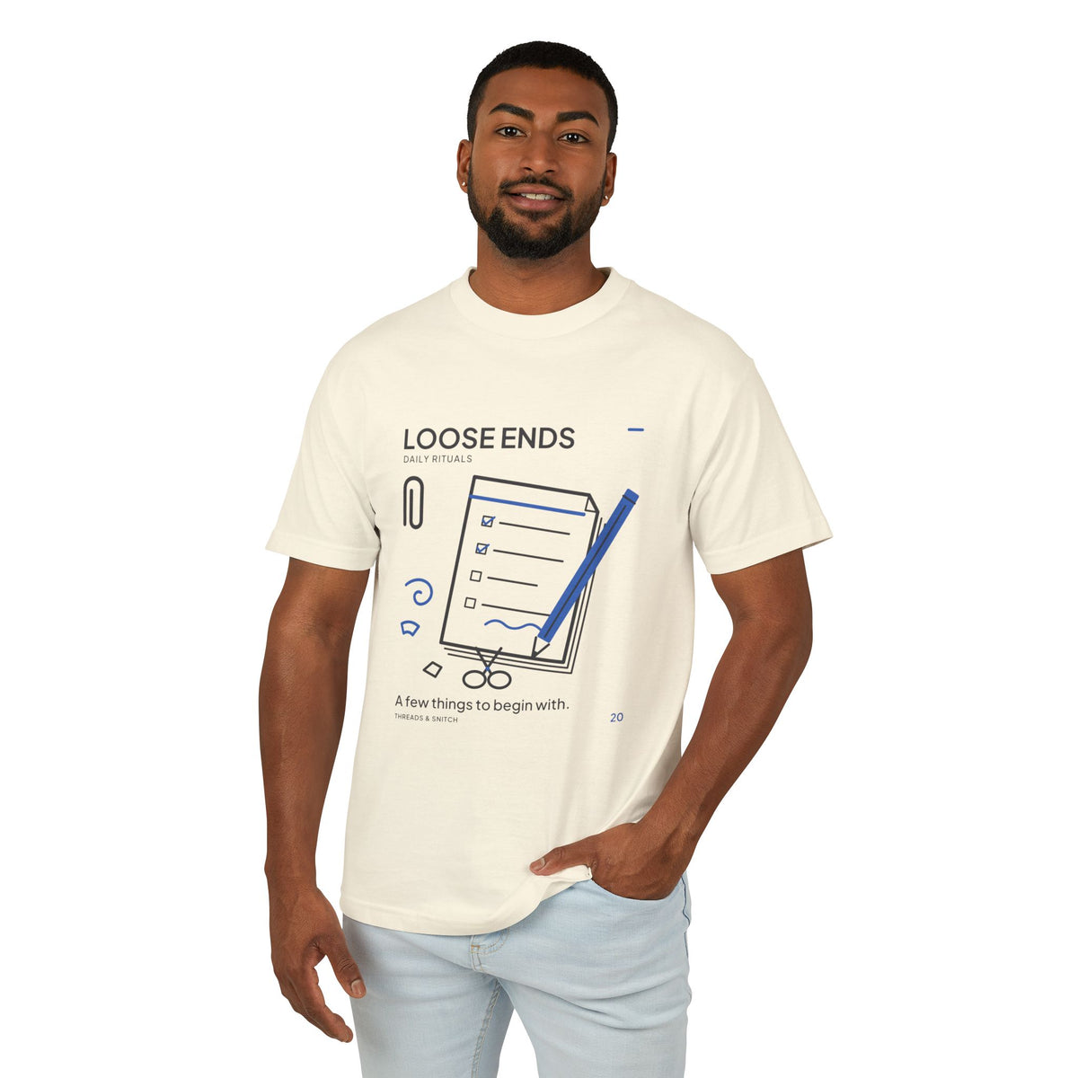 Model mockup of Loose Ends Stationery Tee in Faded Cream, front view showing diagonal pencil over a task pad with scissors and a paper clip.