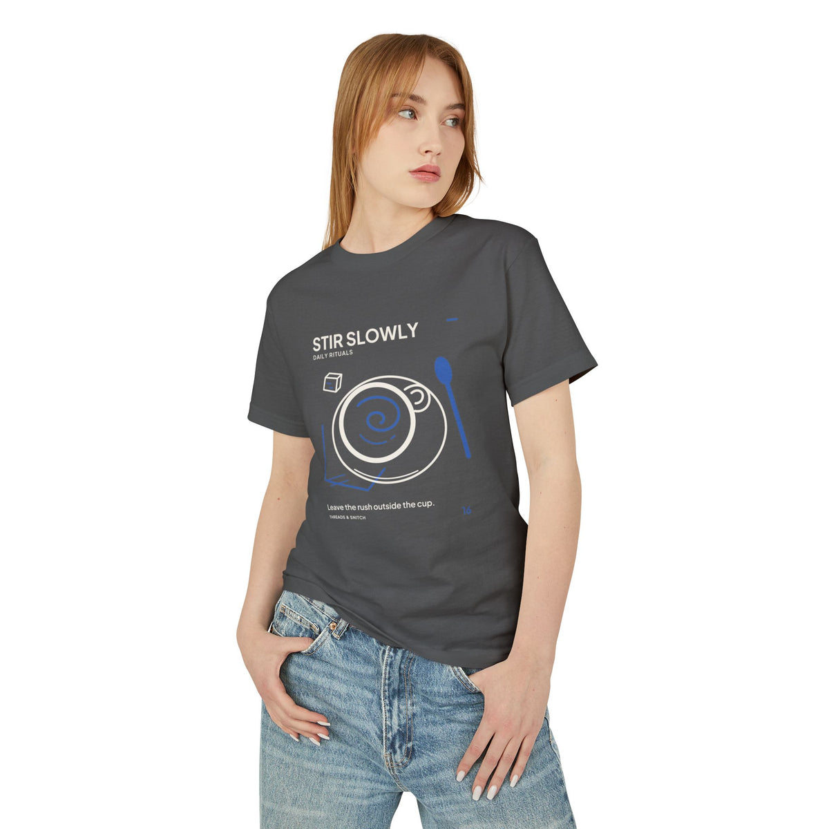 Model mockup of Stir Slowly Cup & Spoon Tee in Faded Black, front view showing cup and saucer with blue stirring spiral and oversized spoon.