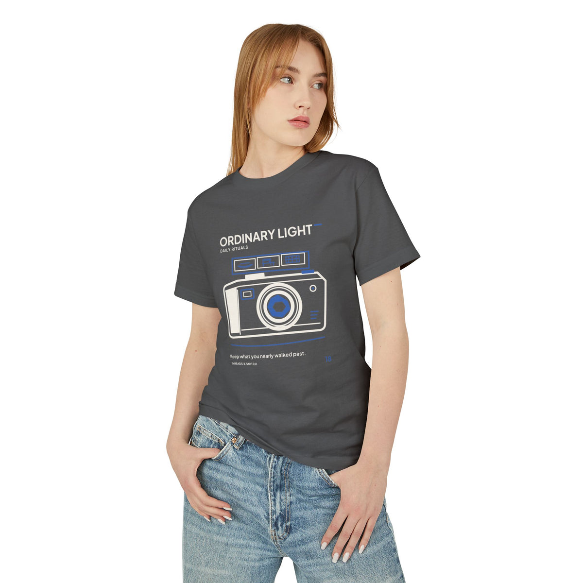 Model mockup of Ordinary Light Camera Tee in Faded Black, front view showing camera with blue lens details and a strip of small domestic scenes.