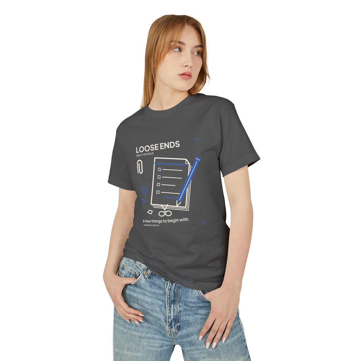 Model mockup of Loose Ends Stationery Tee in Faded Black, front view showing diagonal pencil over a task pad with scissors and a paper clip.