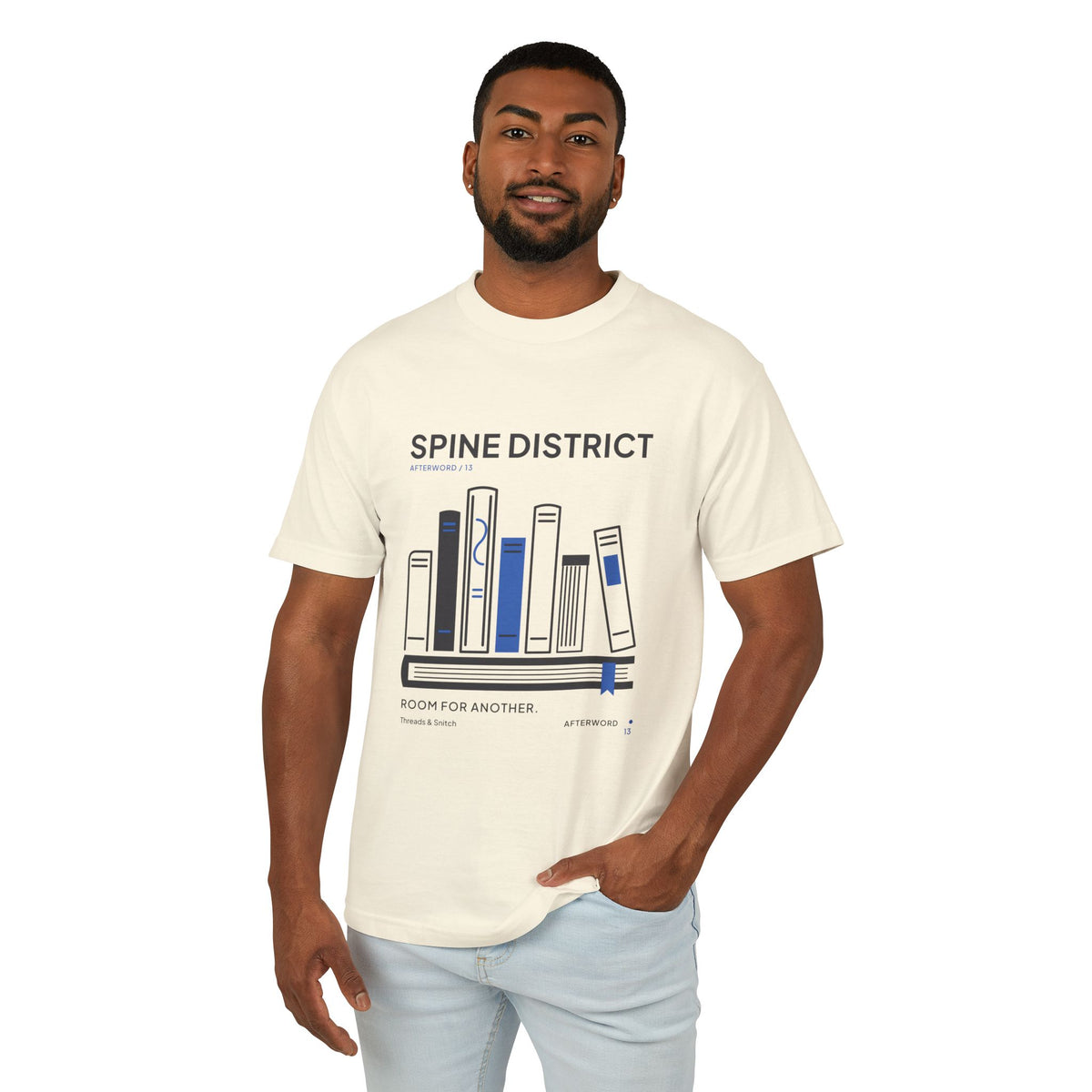 Model mockup of Spine District Book Spine Tee in Faded Cream, front view showing varied book spines arranged as a skyline above a horizontal book.